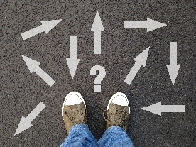Pair of sneakers on asphalt surrounded by many white arrows pointing in different directions, with a central question mark.